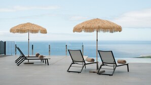Deux fauteuils de jardin dotés de parasols en chaume, installés sur une terrasse offrant une vue sur la mer.