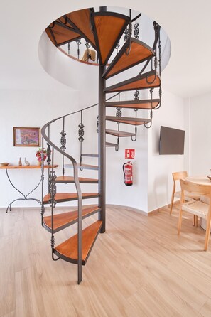 Eine Wendeltreppe mit Holzstufen und Metallgeländern in einem Raum mit Holzboden, einem Tisch mit Stühlen und einem an der Wand befestigten Feuerlöscher.