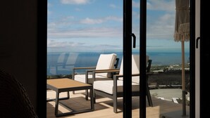 Un balcon avec deux fauteuils blancs dotés de coussins et une table en bois, offrant une vue sur un paysage côtier sous un ciel dégagé.