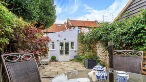 A patio with a round glass table, two chairs, and a white door leading to a house with a red-tiled roof. - Rose Cottage - sleeps 4 guests  in 2 bedrooms (Burnham Market)