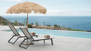 Zona junto a la piscina con dos sillones de playa y un paraguas de paja, con vista a un mar azul y despejado.