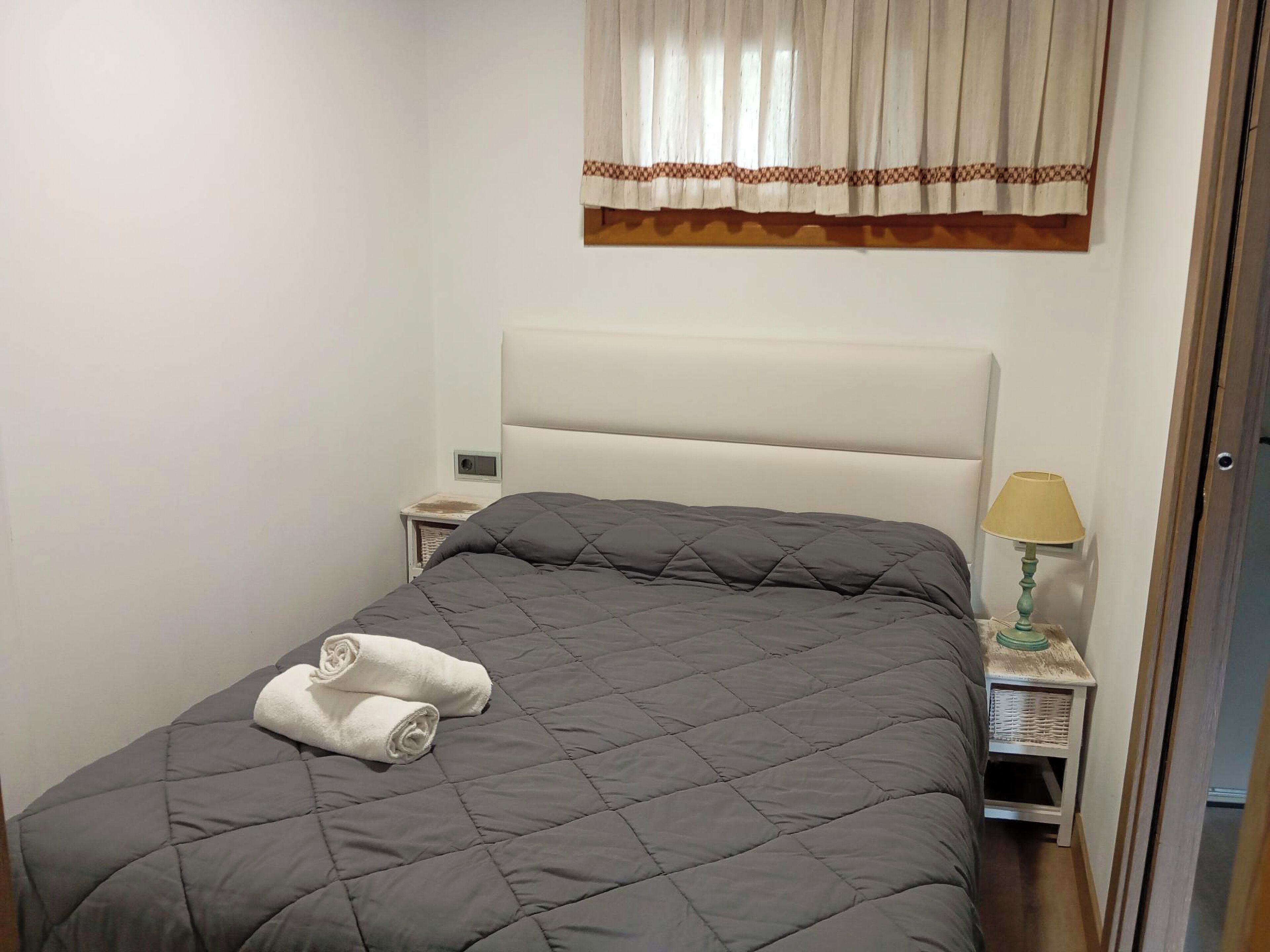 A neatly made bed with a grey quilt, two rolled towels, a bedside lamp, and a window with a curtain.