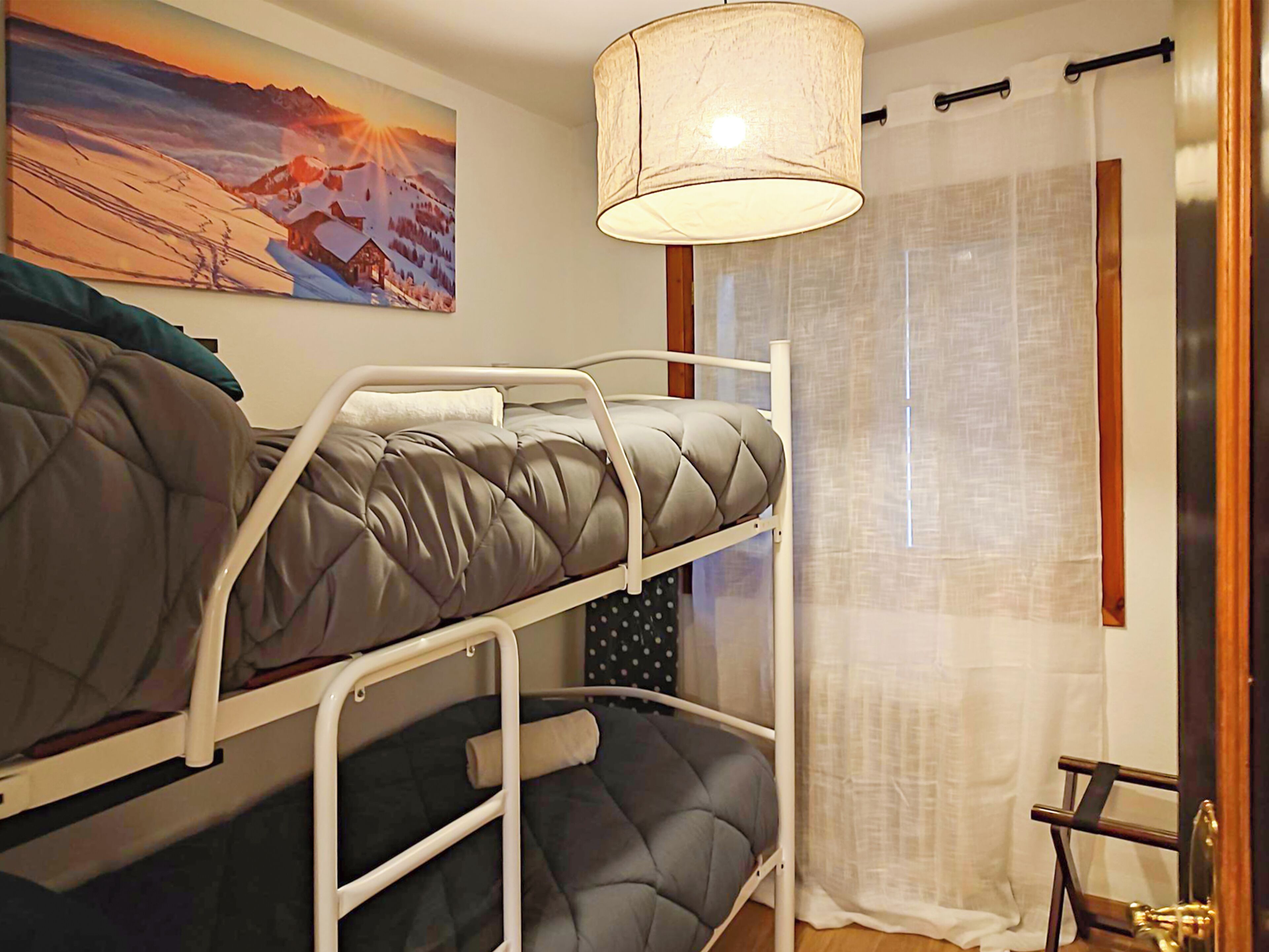 Habitación con literas, un cuadro de un paisaje montañoso nevado, una lámpara colgante y una ventana con cortina.