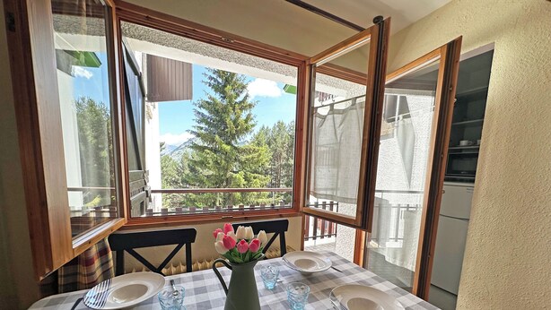 Un coin repas avec une nappe à carreaux, un vase rempli de fleurs et une vue sur les arbres et les montagnes, visible à travers de grandes fenêtres.