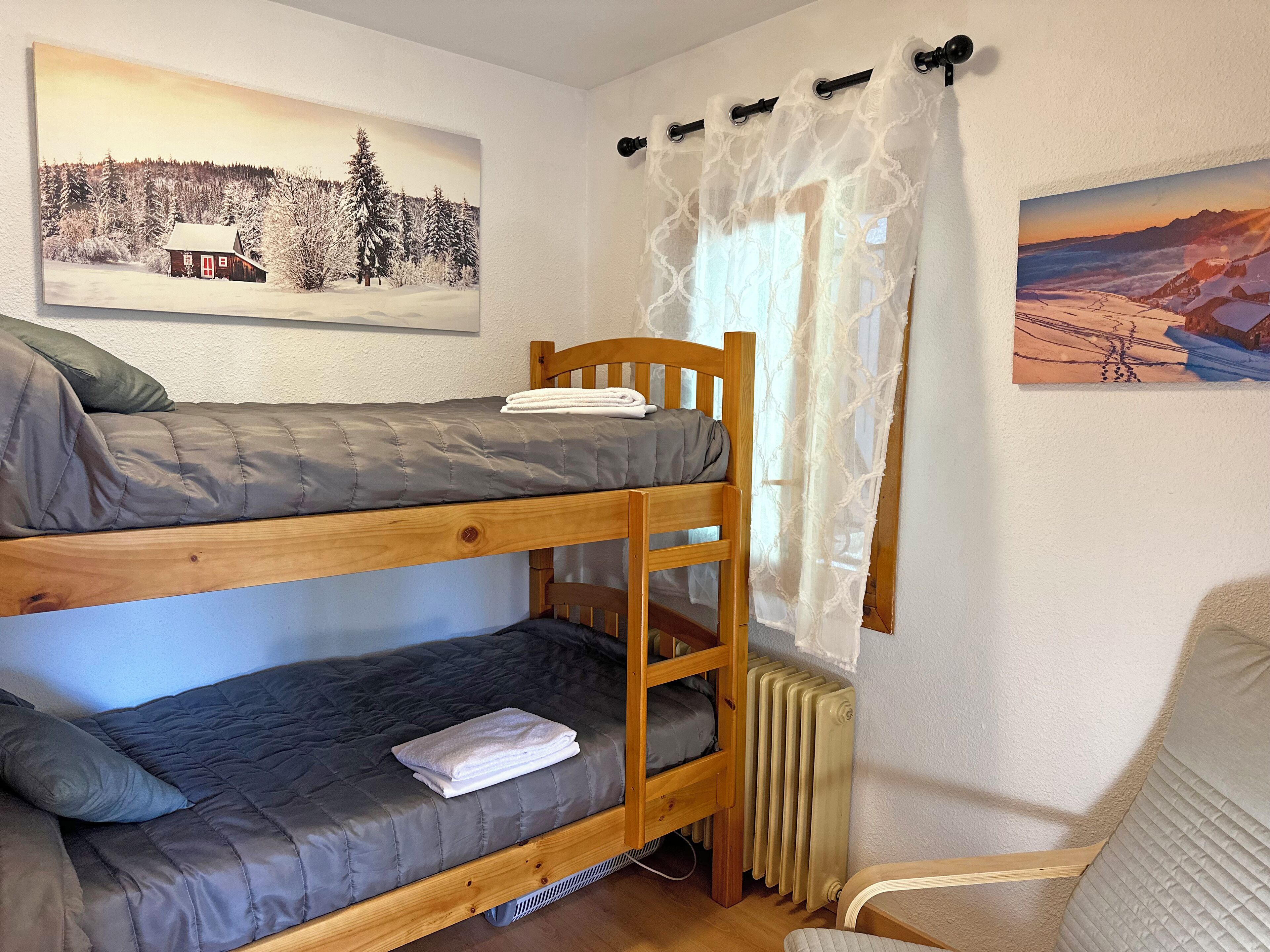 A bunk bed room with a radiator, a wooden chair, and two paintings on the walls.