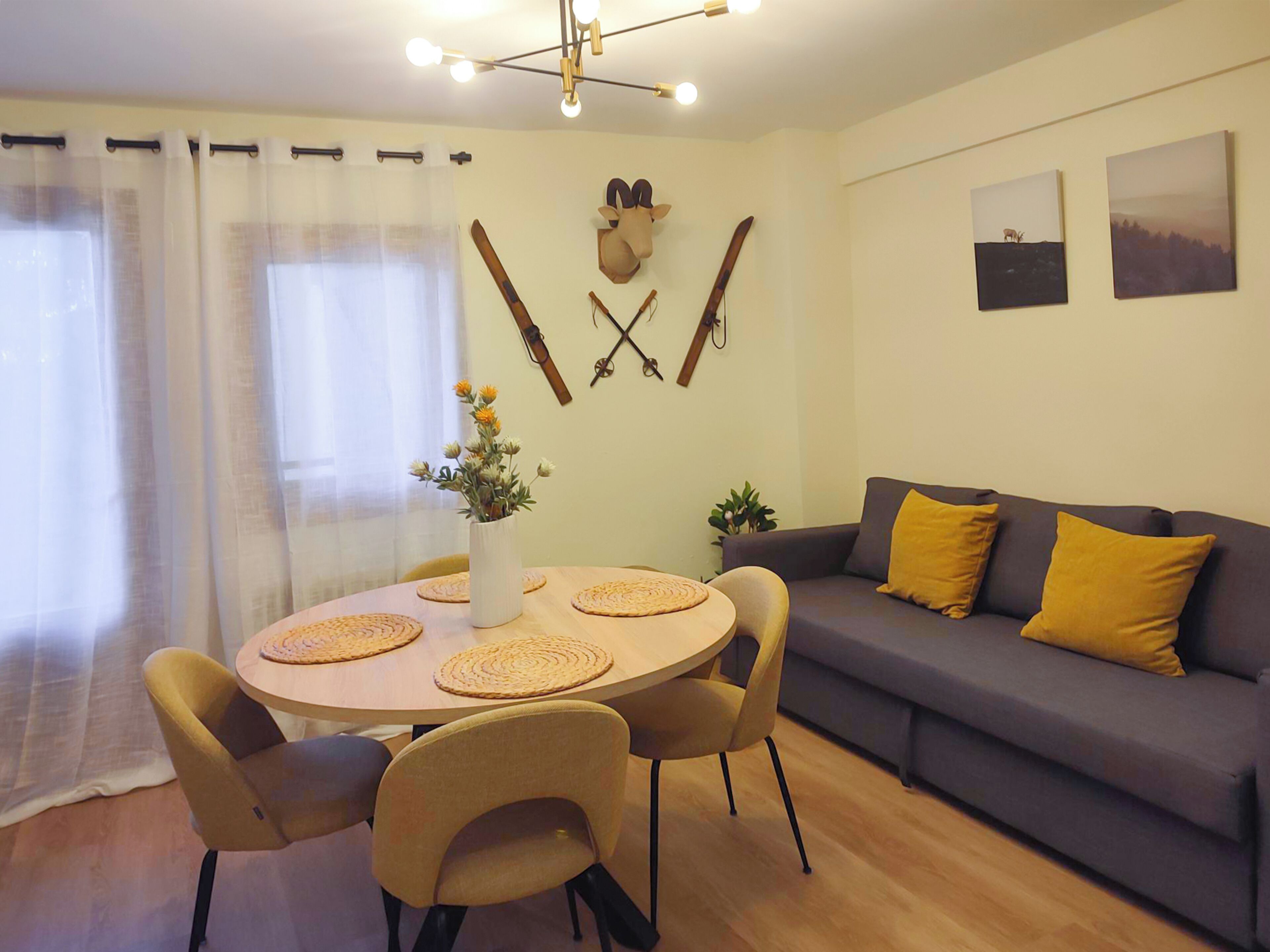 Un coin repas avec une table ronde, des chaises et un vase de fleurs. On y trouve également un canapé avec des coussins jaunes et un porte-manteau mural où sont accrochés des paires de skis et un chapeau.