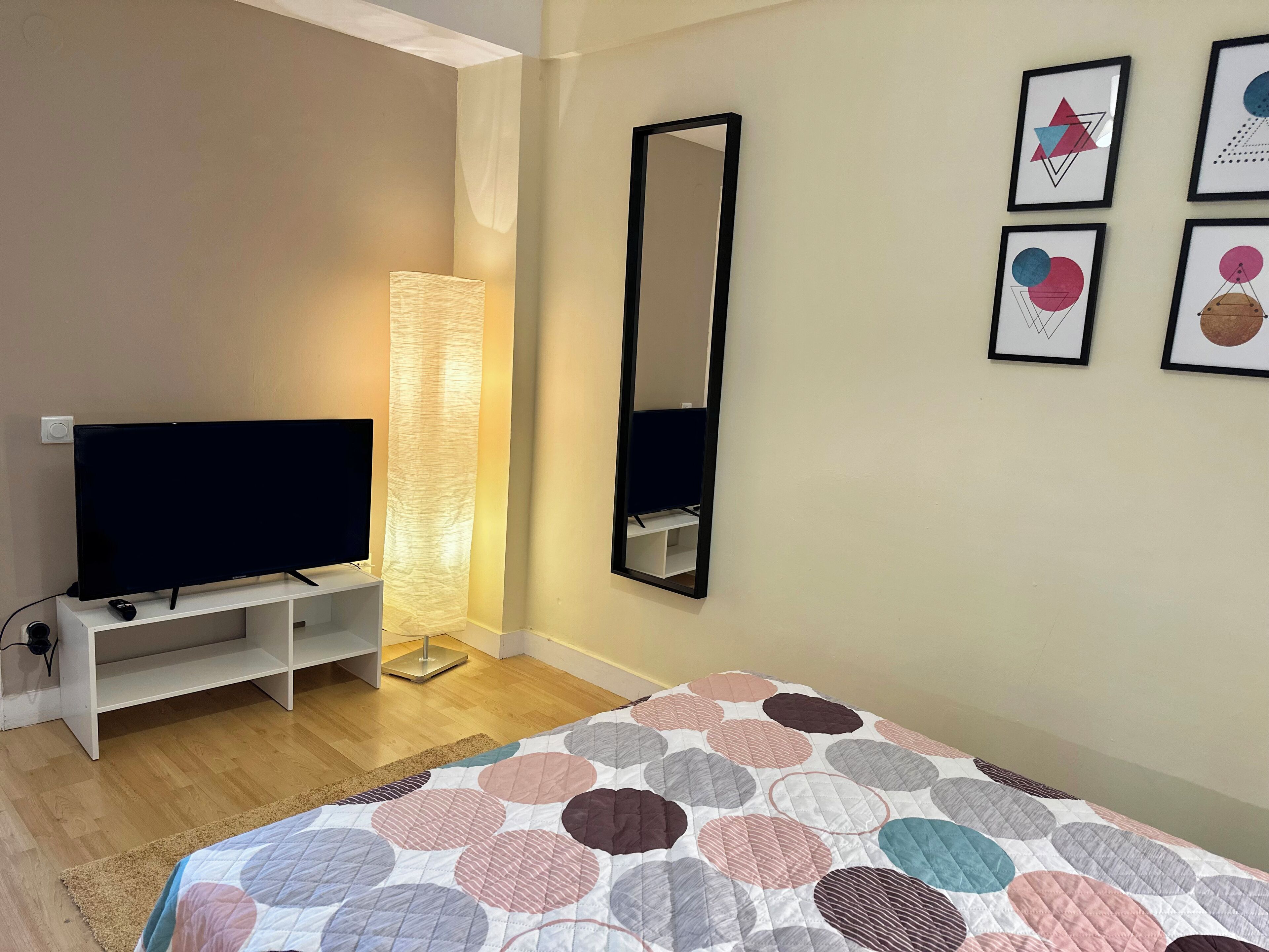 Quarto com televisão, luminária de chão, cama com colcha estampada e obras de arte nas paredes.