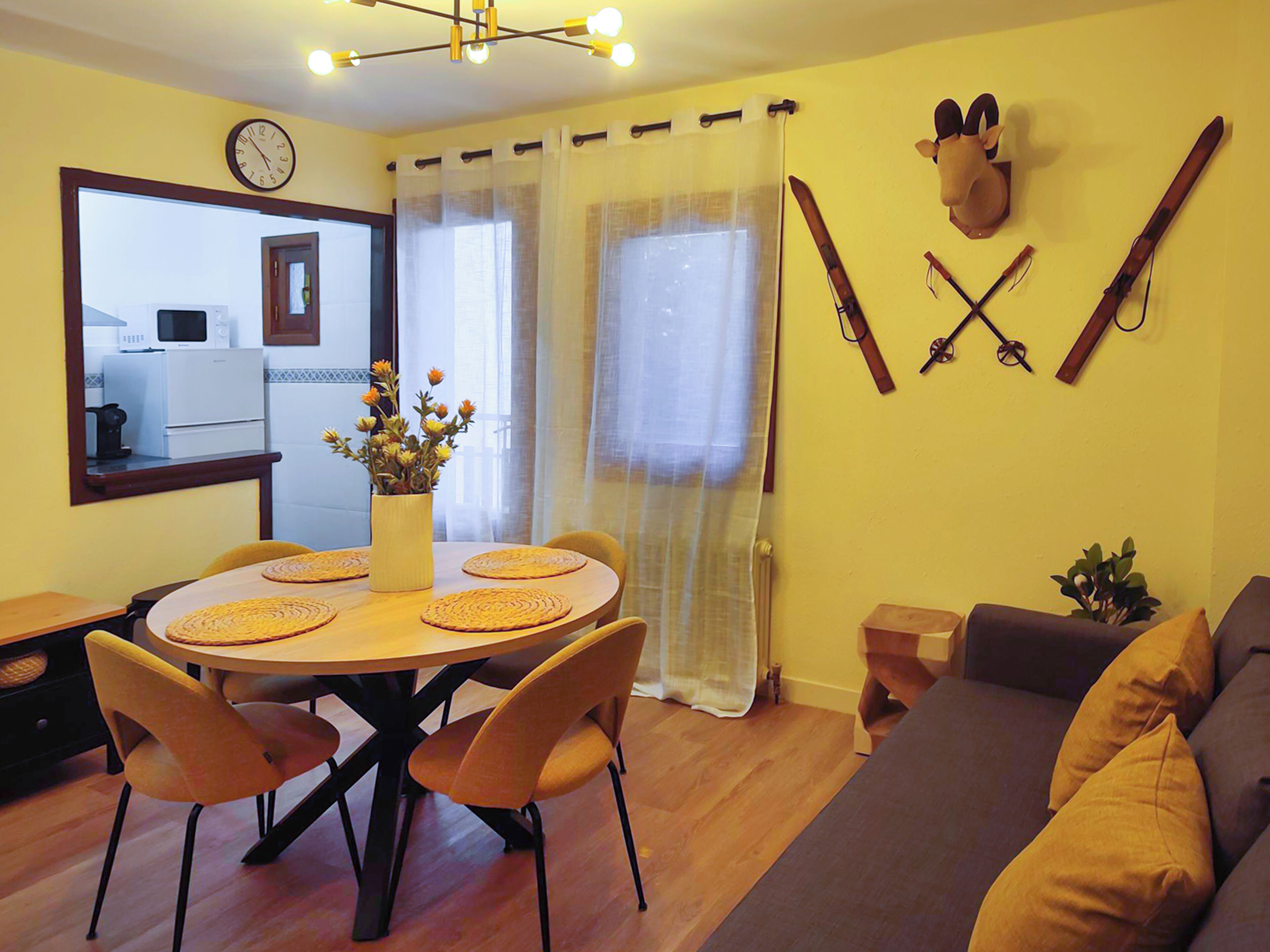 Ein Essbereich mit einem runden Tisch, Stühlen und einem Blumenstrauß. Im Hintergrund ist eine Küche mit einer Mikrowelle und einem Kühlschrank erkennbar.