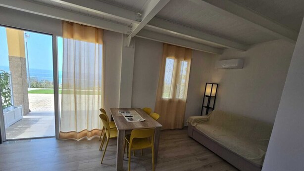 A room with a wooden floor, a dining table with yellow chairs, a sofa, and a window with curtains. - RIVIELLO ROOMS. (Marina di Camerota)