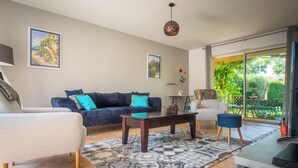 A spacious living room with a sofa, armchair, and coffee table, a dining area with a table and chairs, and a large sliding glass door leading to a garden. - La Bienvenue - Four Bedroom Villa, Sleeps 8 (Pézenas)