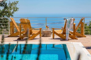 Un espace aménagé au bord de la piscine, avec des chaises et une table en bois, offrant une vue sur une mer d’un bleu limpide et sur des montagnes lointaines.