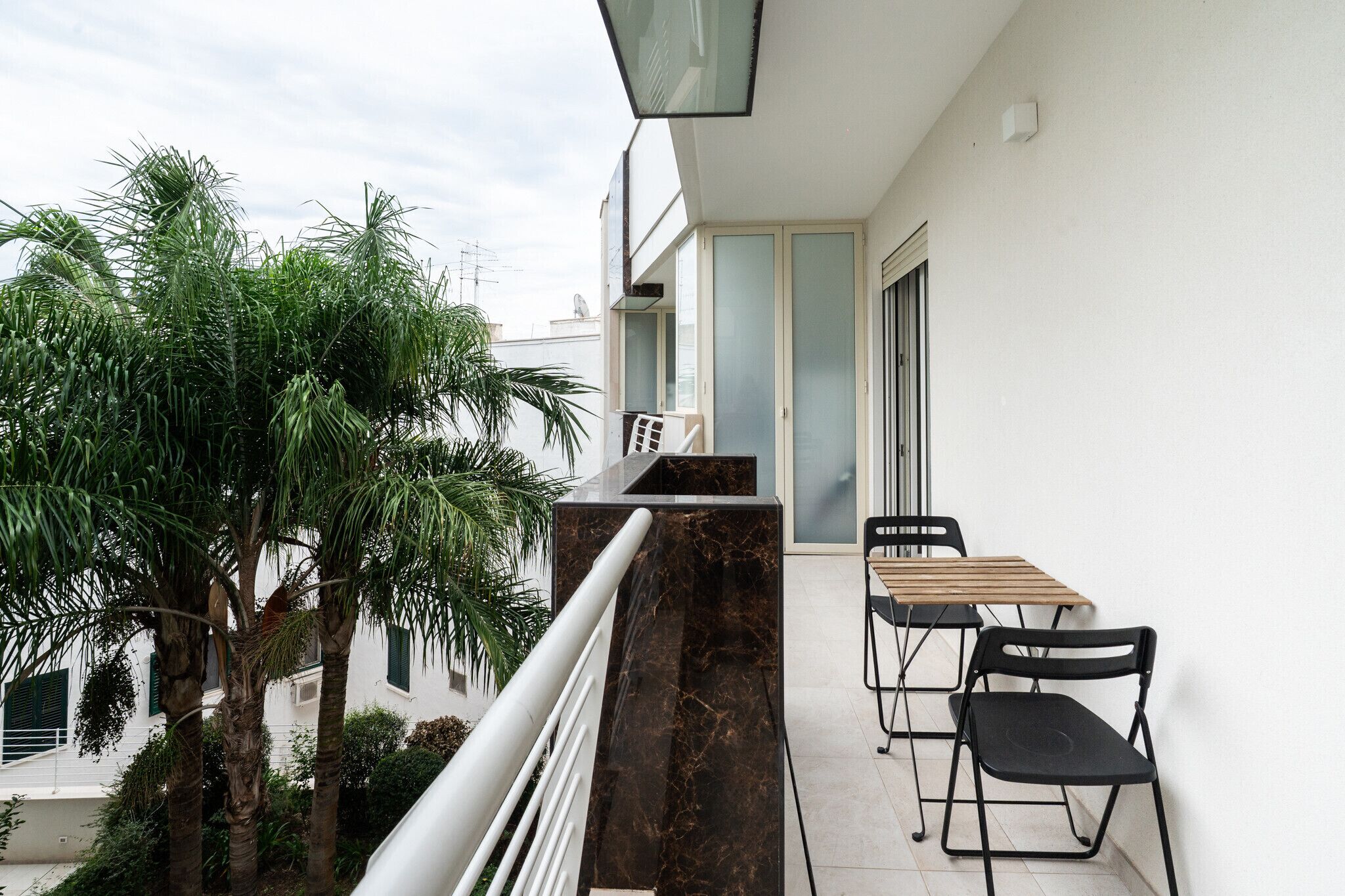 A balcony with a marble column, a wooden table, two black chairs, and a potted palm tree.