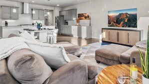 En moderne stue med sofa, skammel og sofabord. I baggrunden er der et køkken med grå skabe og et spiseområde.
