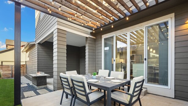 En udendørs patio med et bord og stole, en pergola med lyskæder, og en skydedør, der fører ind i et rum.