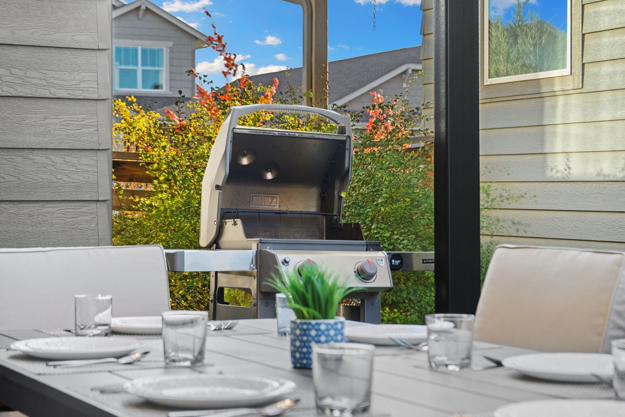 An outdoor dining area with a grill, table set with plates and glasses, and a potted plant.