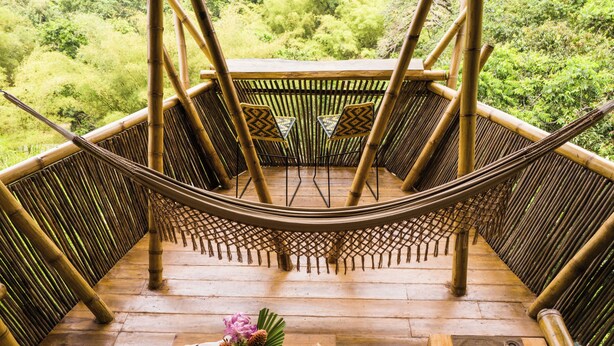 Une terrasse en bois avec un hamac, des meubles en bambou, une table ornée d’un vase de fleurs et d’un livre.