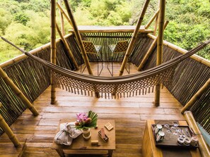 A wooden deck with a hammock, bamboo furniture, and a table with a vase of flowers and a book.