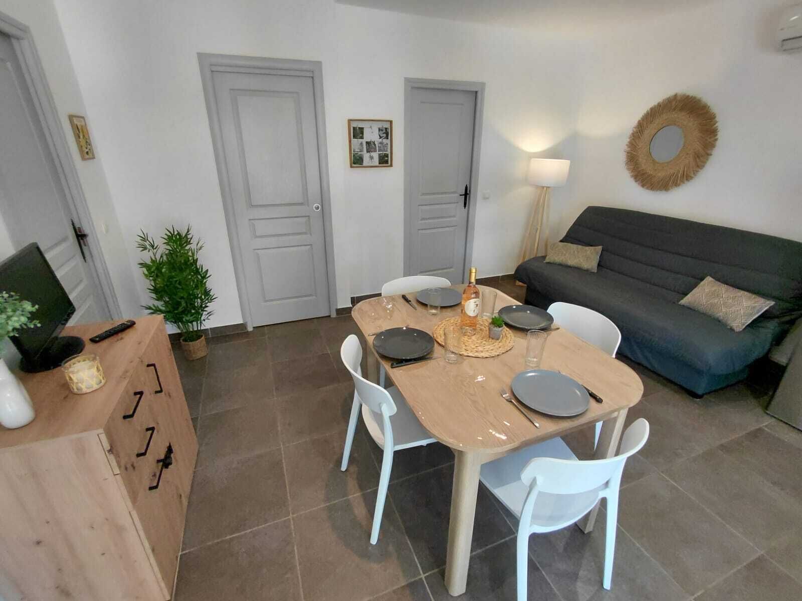 Un comedor con una mesa lista para cuatro, sillas blancas, un sofá gris, una lámpara y una planta pequeña.