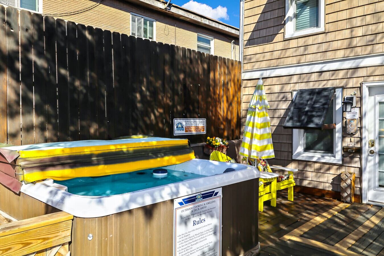 A backyard with a hot tub, a wooden deck, a yellow umbrella, and a house with a window and a door.
