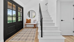Un interior moderno con una puerta negra, escalera blanca y un banco con un espejo y una bolsa que dice 'HELLO WEEKEND'.