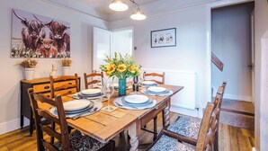 Een eetkamer met een houten tafel gedekt voor vier personen, omringd door houten stoelen. Op de tafel staat een vaas met zonnebloemen, en aan de muur hangt een schilderij van jakken.