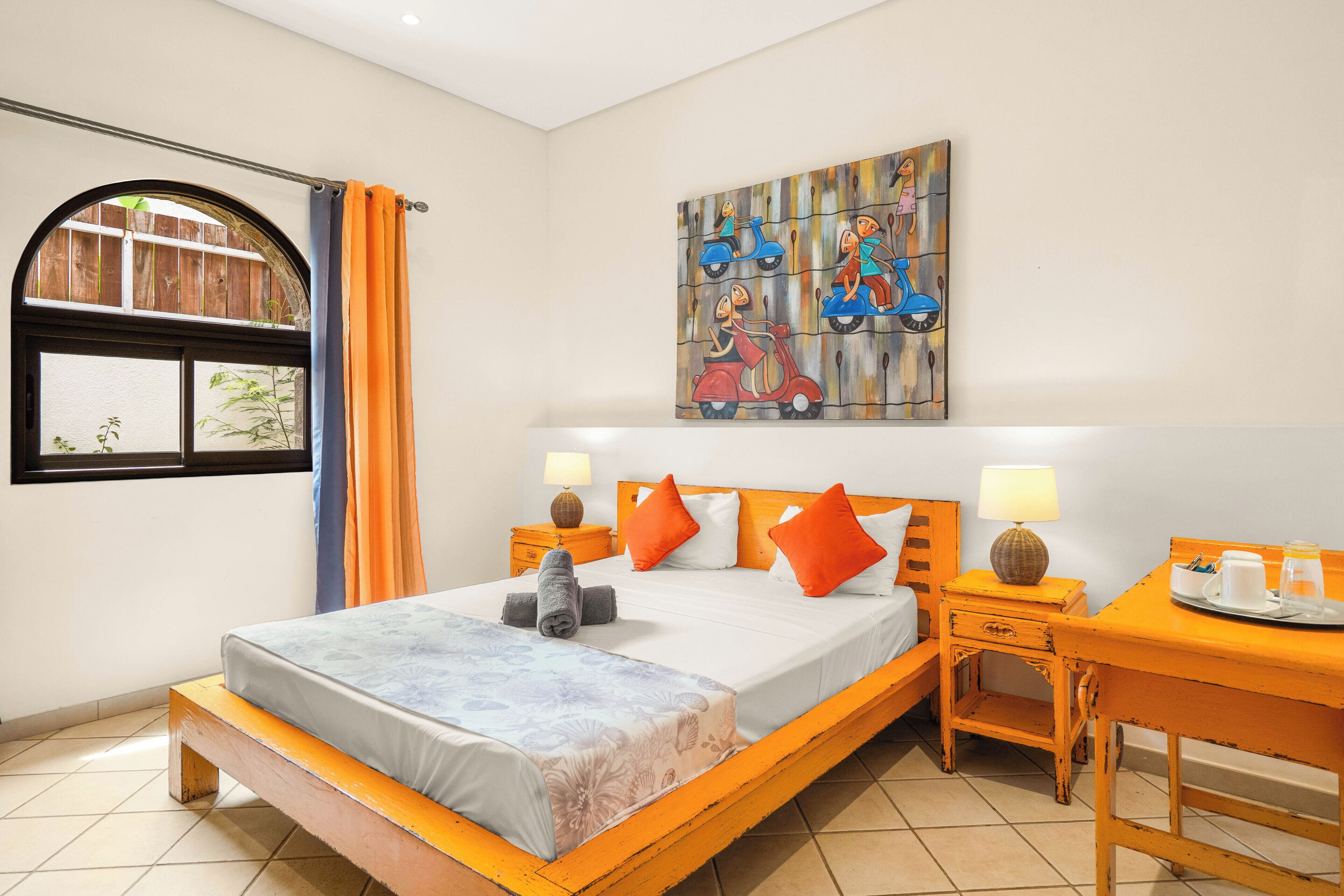 A bedroom with a bed, a nightstand, a desk, a window with curtains, and a wall painting.