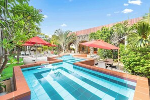 A swimming pool with tiled edges, surrounded by lounge chairs and red umbrellas, set against a backdrop of greenery and stone walls. - Aroma Plaza Boutique Hotel (Cap Malheureux)