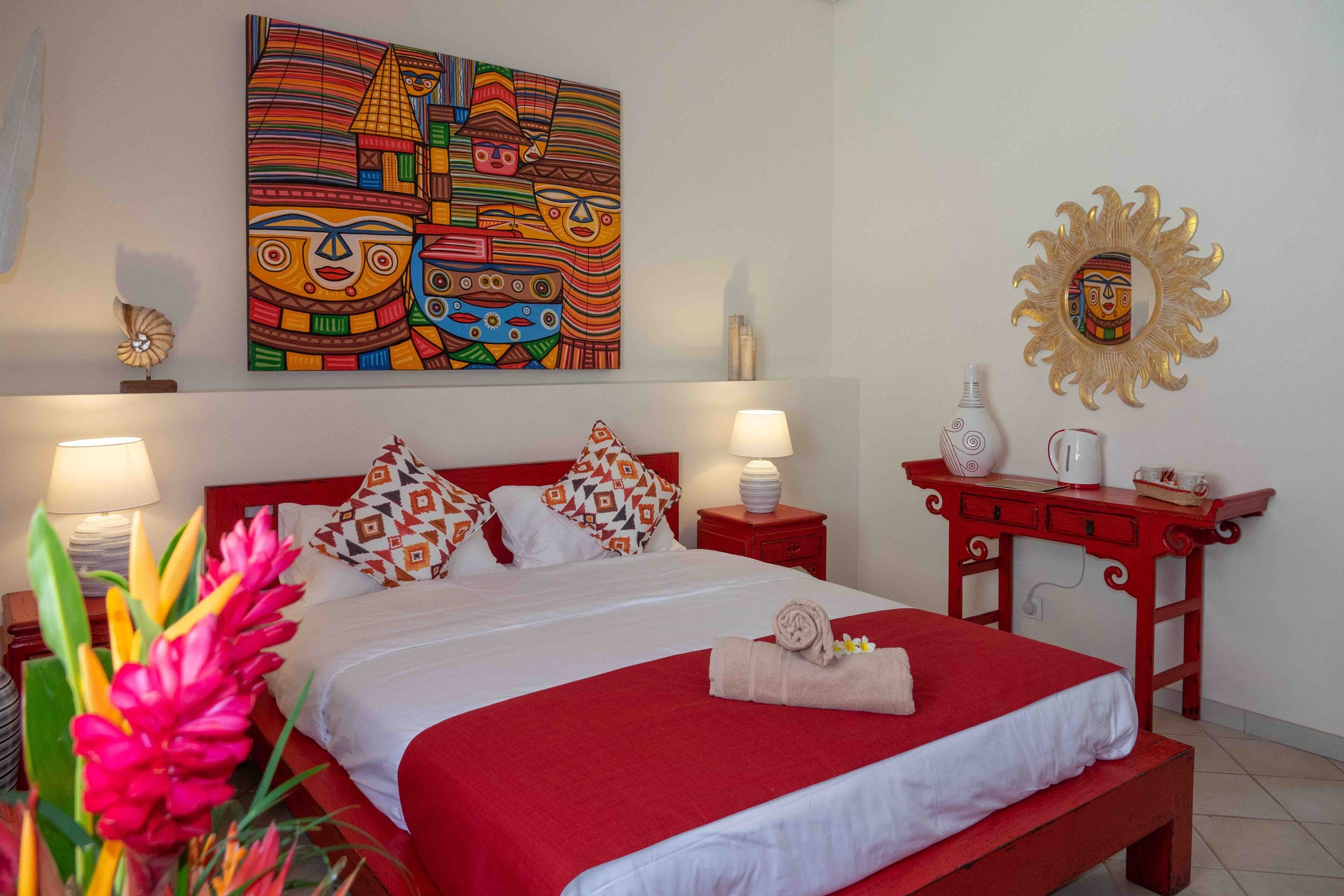 Quarto de hotel com uma cama grande, roupa de cama vermelha e branca, uma obra de arte colorida na parede, um espelho decorativo e uma mesa de cabeceira vermelha com uma luminária.