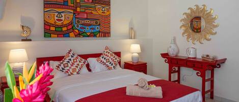 Quarto de hotel com uma cama grande, roupa de cama vermelha e branca, uma obra de arte colorida na parede, um espelho decorativo e uma mesa de cabeceira vermelha com uma luminária.