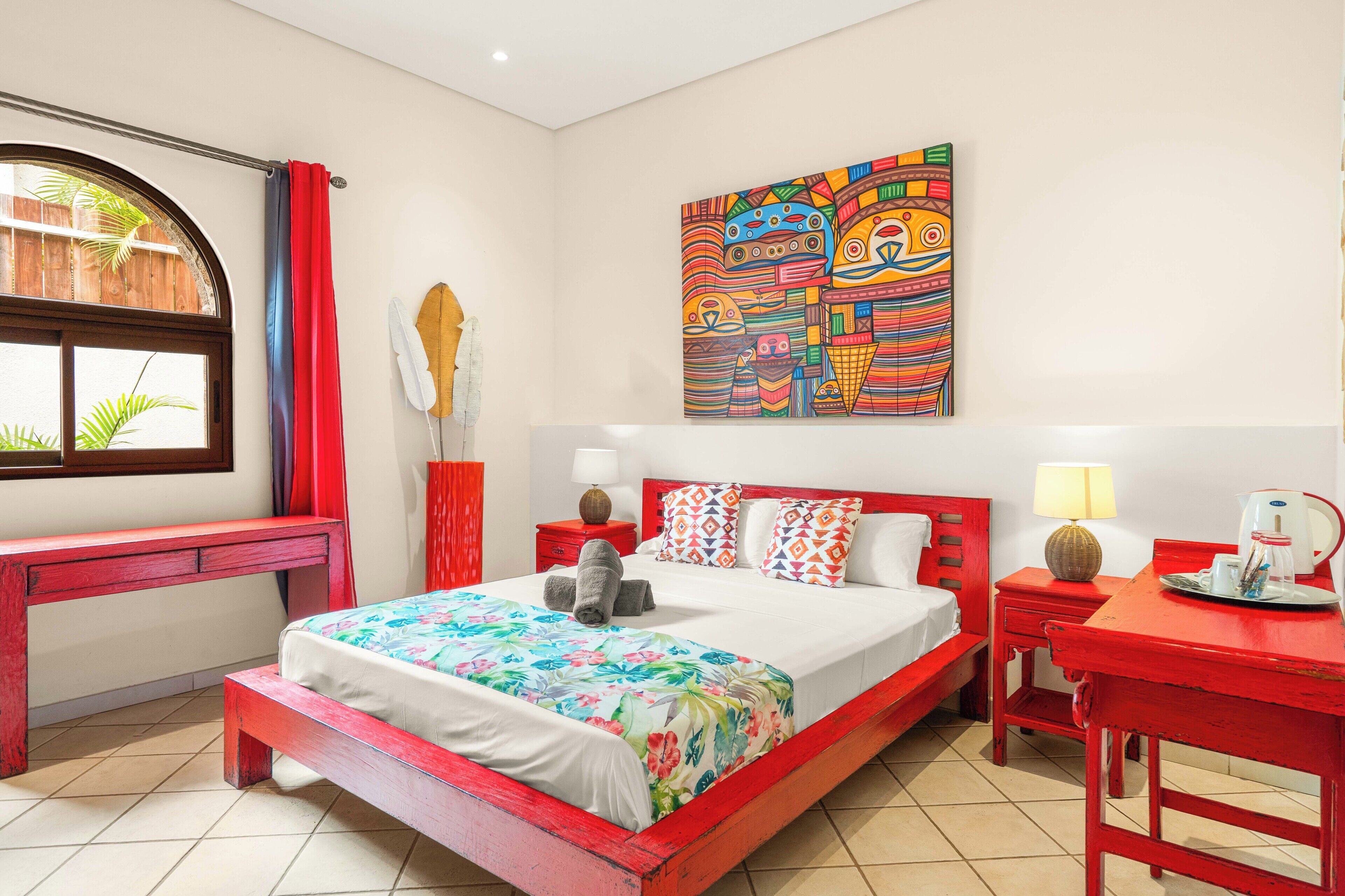 Een kamer met een bed, een bureau, een stoel, een raam met gordijnen, een wanddecoratie en een nachtkastje met een lamp en een waterkoker.