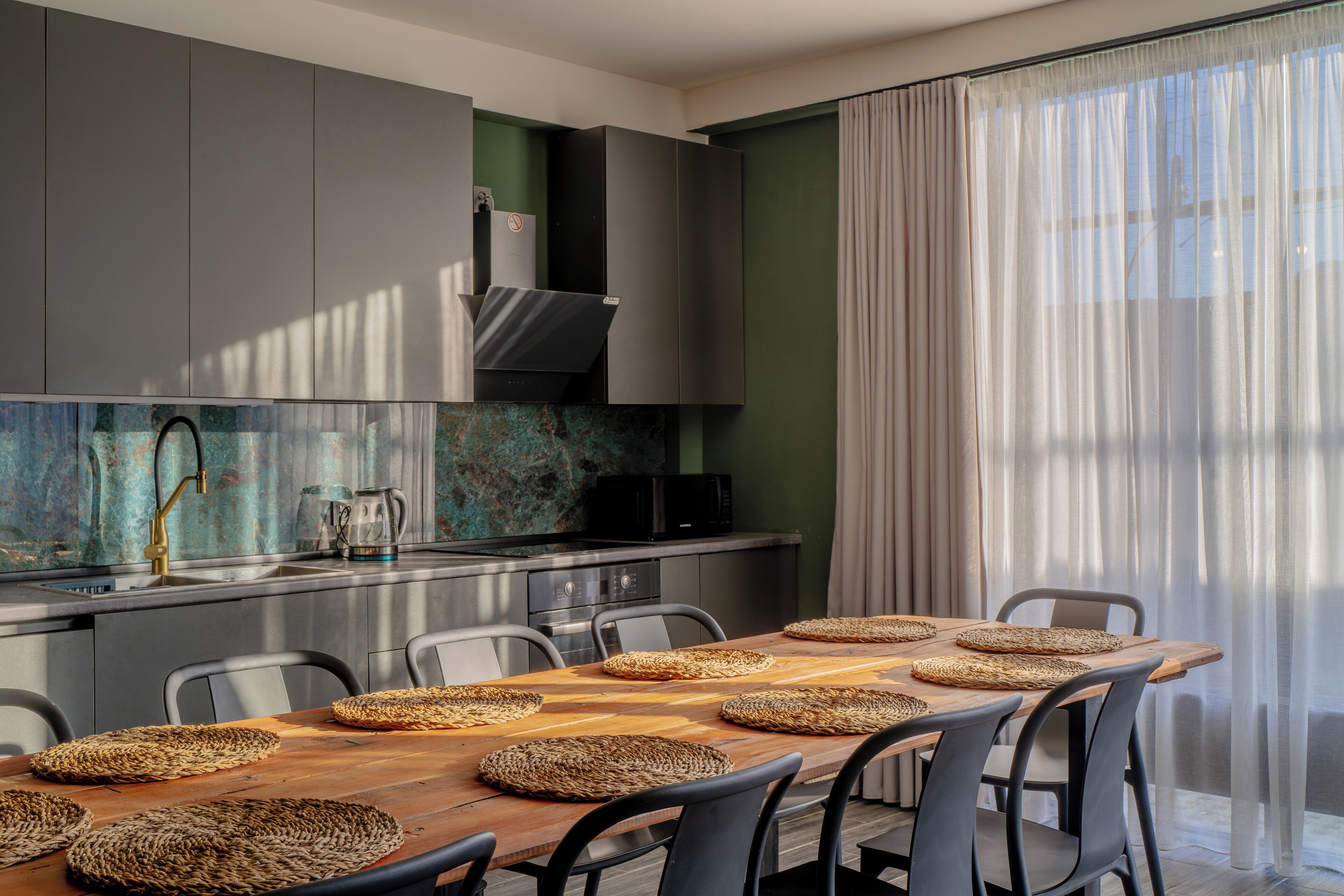 A modern kitchen with a wooden dining table, grey cabinets, and a large window with sheer curtains.