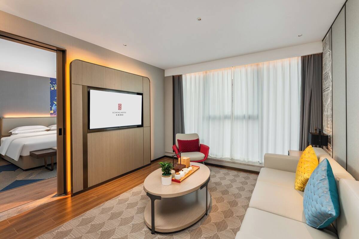 Een moderne hotelkamer met een bed, een bank, een salontafel en een televisiescherm met hotelpromotie.