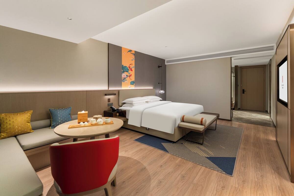 Een moderne hotelkamer met een bed, een bank, een rode stoel, een klein tafeltje met dienblad en een groot raam.