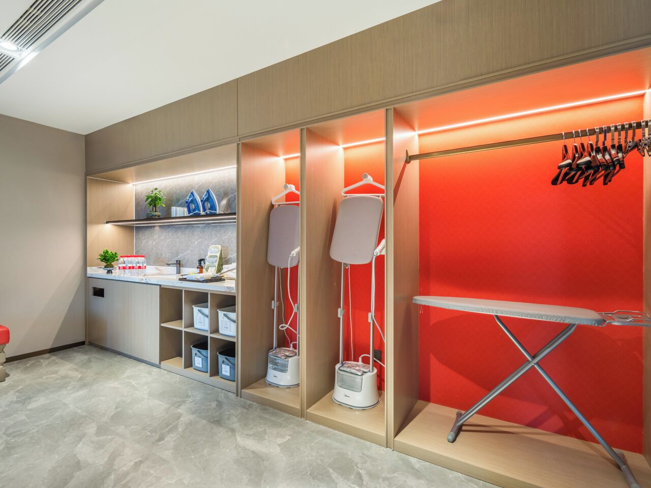 Habitación de hotel moderna con suelo de madera, un escritorio con computadora y planta, dos lámparas de pie y una pared de acento roja.