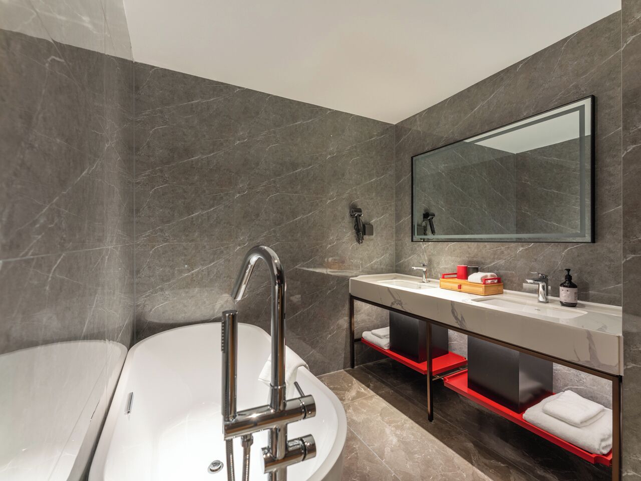 A modern bathroom with a freestanding bathtub, a large mirror, and a vanity with a red shelf.