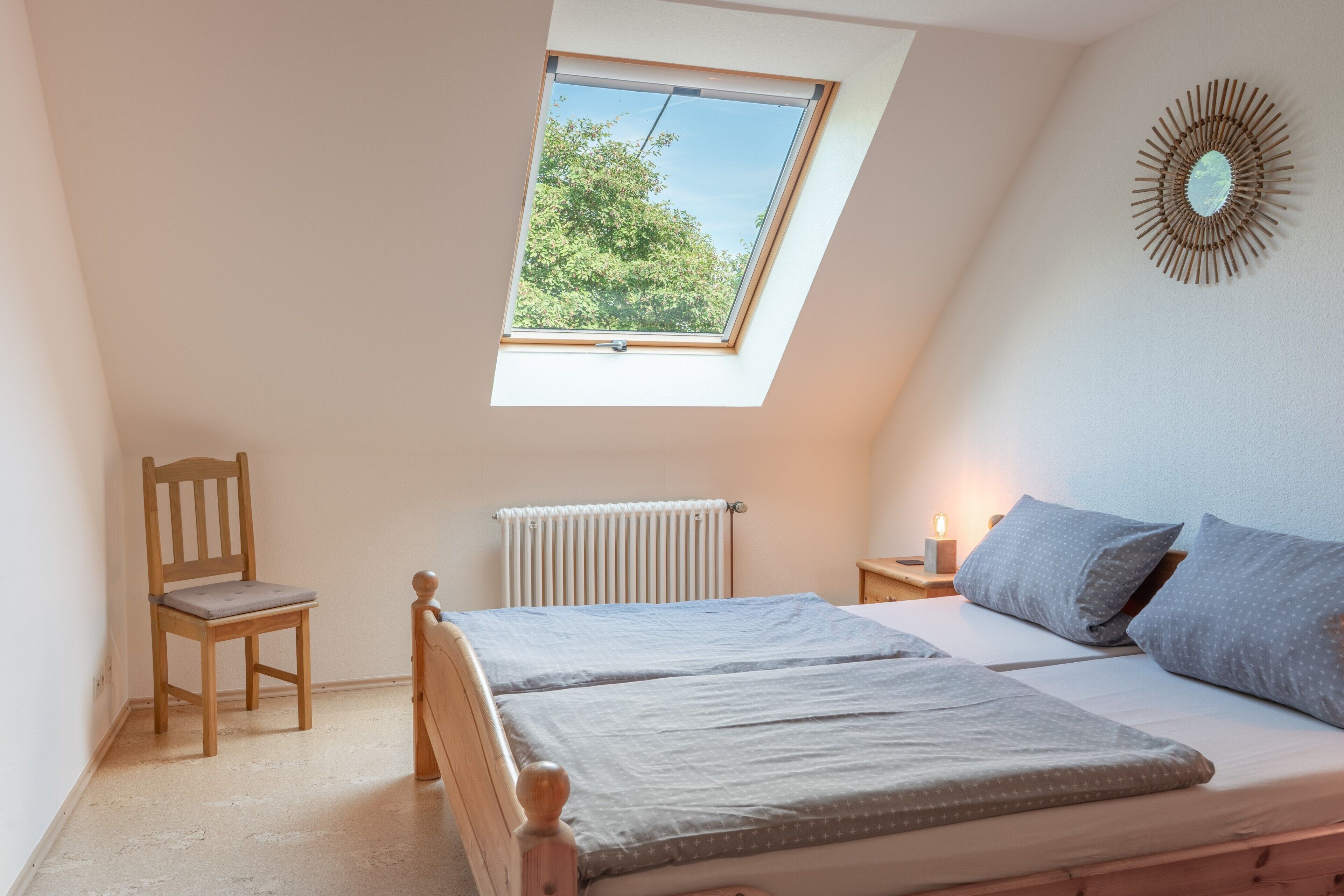 Een slaapkamer met dakraam, een houten stoel, een bed met grijze beddengoed en een nachtkastje met lamp.