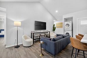 Ein modernes Wohnzimmer mit einer Couch, Sesseln, einem Fernseher, einem Couchtisch und einer Waschmaschine.