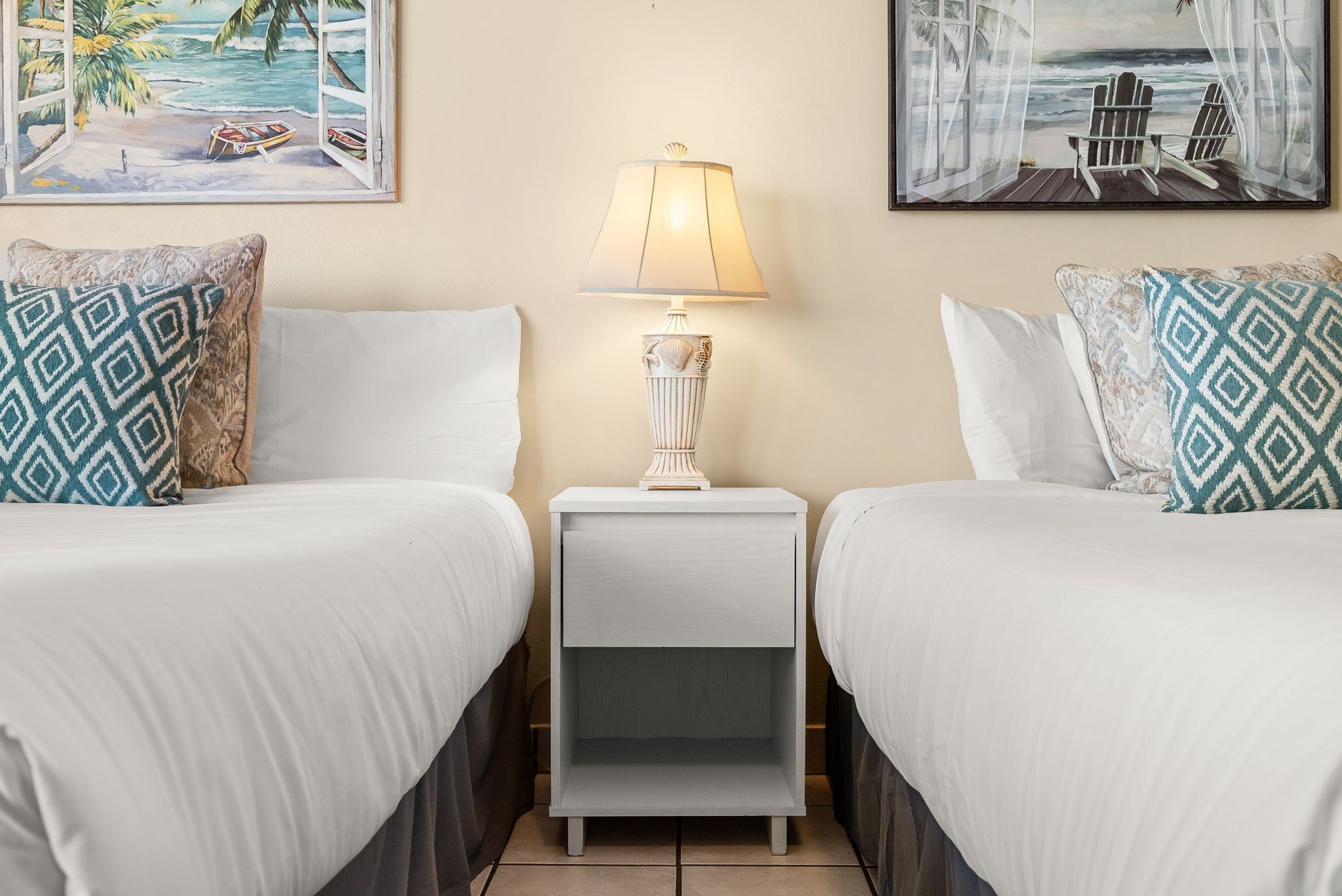 Een hotelkamer met twee bedden, een nachtkastje met een lamp, en twee ingelijste strandtaferelen aan de muur.