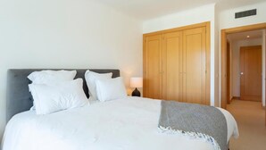 Una cama bien hecha con sábanas y almohadas blancas, una manta gris y un armario de madera al fondo.