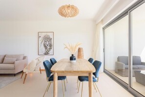 A modern living room with a wooden dining table, blue chairs, a white sofa, a large window, and a ceiling light fixture. - Snazzy Varandas do Castelo One Bedroom Apartment Sleeps 2 Persons (Albufeira)