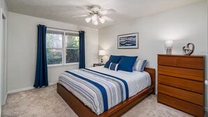 A bedroom with a bed, a wooden dresser, a ceiling fan, a window with curtains, and a painting on the wall.