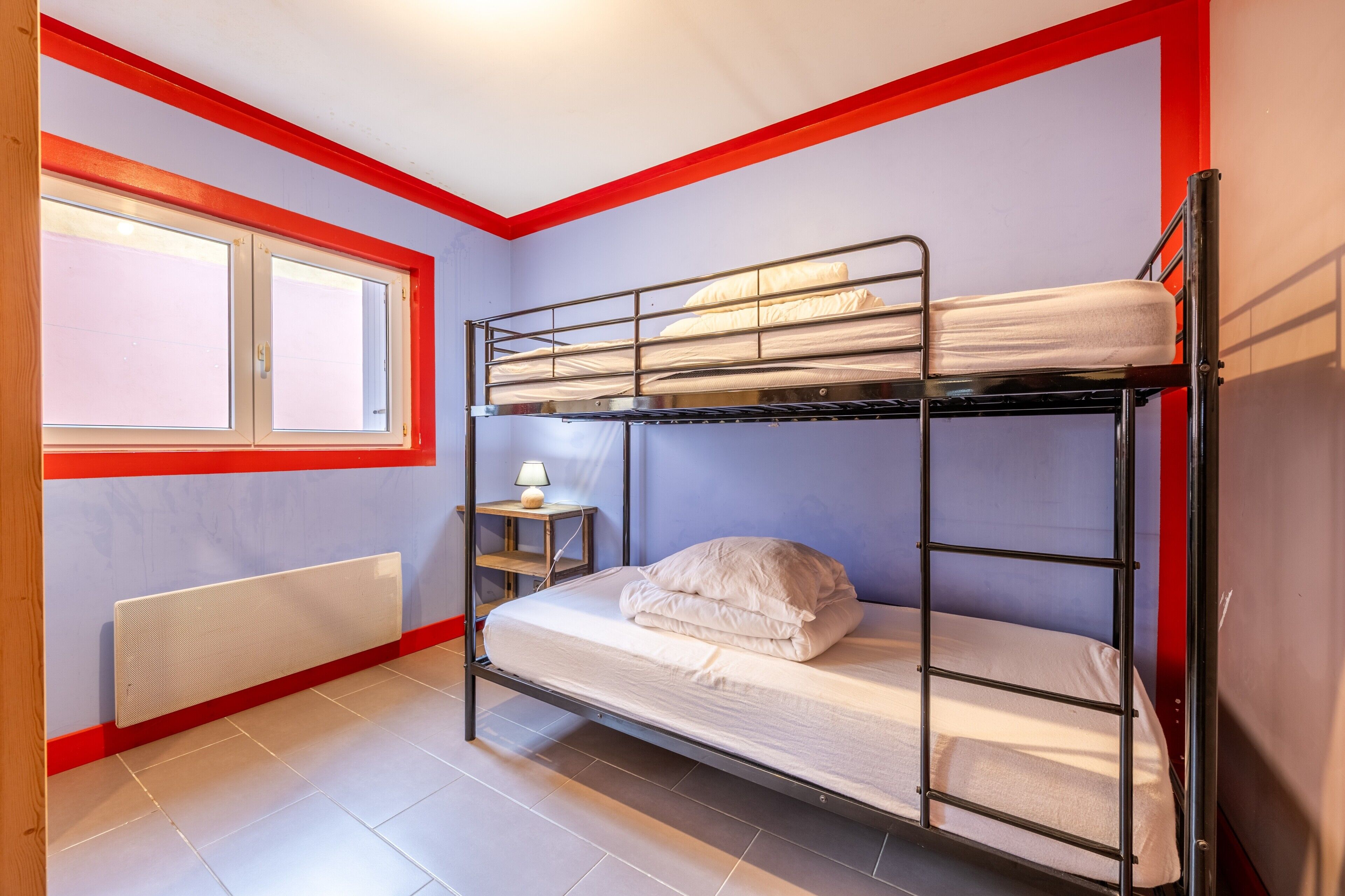 A bunk bed room with a window, a radiator, and a small table with a lamp.