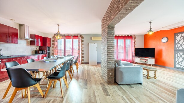 A modern kitchen with red cabinets, a dining area with a long table and chairs, and a living room with a TV and seating. - Holiday Home 'Sleeps 22 Brem Center with Large' (Brem-sur-Mer)