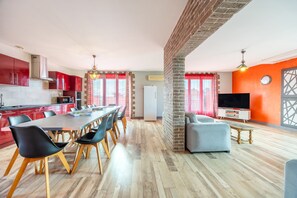 A modern kitchen with red cabinets, a dining area with a long table and chairs, and a living room with a TV and seating.