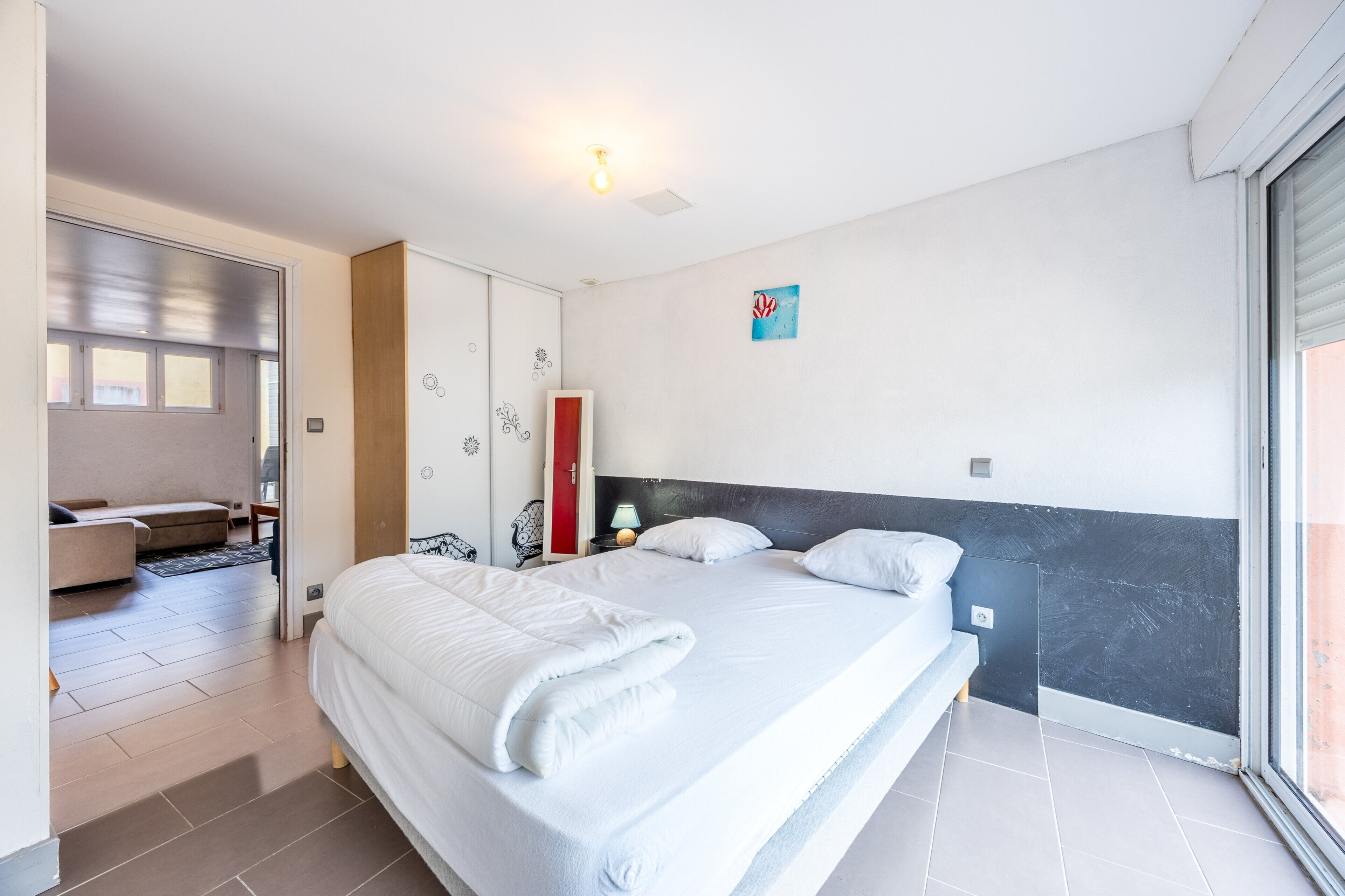 Ein modernes Schlafzimmer mit einem großen Bett, einem Kleiderschrank, einem Nachttisch mit Lampe und einem Fenster.