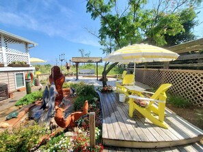 Une terrasse en bois avec des fauteuils Adirondack jaunes, un petit jardin agrémenté d’une sculpture de poisson et une pergola surmontée d’un parasol jaune.