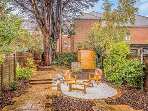 Un jardin avec un chemin de pierres, des chaises en bois, un foyer et un abri de jardin en bois.
