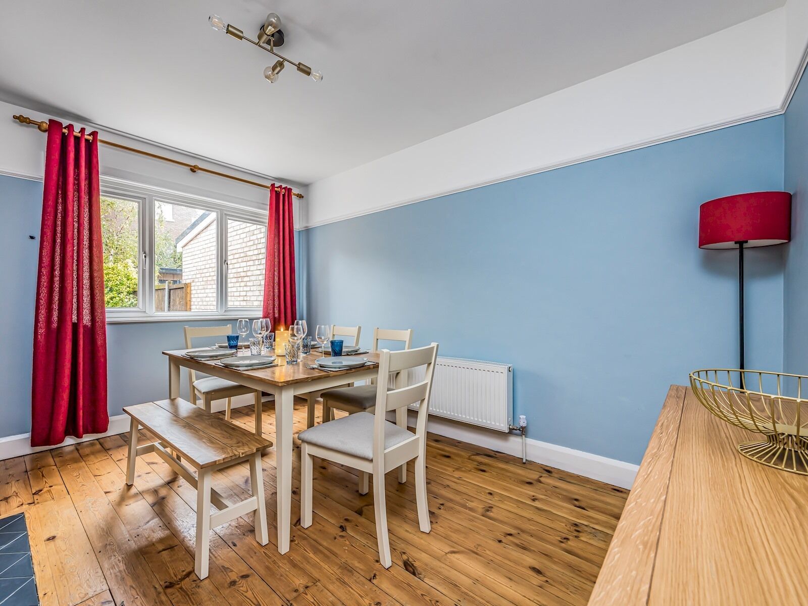 Un coin repas avec une table en bois dressée pour quatre personnes, des chaises blanches, un rideau rouge, un radiateur blanc et un lampadaire rouge.