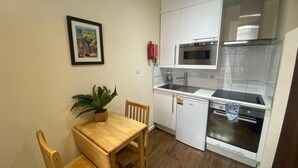 Ett kompakt kök med vita skåp, en mikrovågsugn, en ugn och ett litet kylskåp. Där finns ett matbord i trä med stolar och en krukväxt.