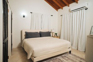 Un dormitorio con cama, cortinas, un equipo de aire acondicionado, una mesita de noche y una lámpara.